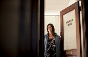 A woman with dark hair is standing at an open door labeled 'American Fed of Musicians.' She is wearing a denim shirt over a patterned top and a necklace, looking directly at the camera with a slight smile.