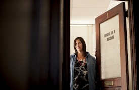 A woman with dark hair is standing at an open door labeled 'American Fed of Musicians.' She is wearing a denim shirt over a patterned top and a necklace, looking directly at the camera with a slight smile.