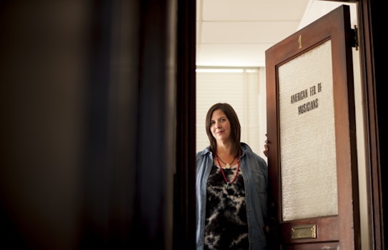 A woman with dark hair is standing at an open door labeled 'American Fed of Musicians.' She is wearing a denim shirt over a patterned top and a necklace, looking directly at the camera with a slight smile.