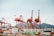 Several large container cranes are positioned at a busy shipping port, with numerous stacked freight containers prominently displaying brand names like Maersk and Evergreen. The backdrop features a mountain range under a clear sky.