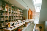 A modern library interior with a high ceiling and natural light filtering through large windows. There are shelves filled with a variety of books along the left side wall and two long wooden tables in the center displaying open books and magazines. The decor includes minimalistic lighting fixtures and a neutral color palette with pops of bright hues from furniture.