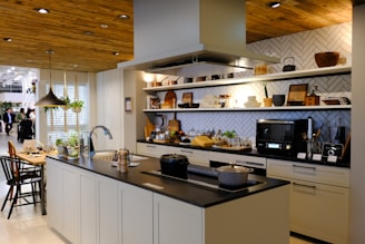 A modern kitchen with a central island featuring a sink and culinary tools. The background includes open shelves displaying various kitchenware such as plates, bowls, and teapots. A microwave is placed on the countertop, and there is a stylish black overhead lamp. The area includes some potted plants, adding a touch of greenery. Wood accents on the ceiling and shelves provide a warm, inviting atmosphere.