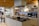 A modern kitchen with a central island featuring a sink and culinary tools. The background includes open shelves displaying various kitchenware such as plates, bowls, and teapots. A microwave is placed on the countertop, and there is a stylish black overhead lamp. The area includes some potted plants, adding a touch of greenery. Wood accents on the ceiling and shelves provide a warm, inviting atmosphere.