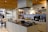 A modern kitchen with a central island featuring a sink and culinary tools. The background includes open shelves displaying various kitchenware such as plates, bowls, and teapots. A microwave is placed on the countertop, and there is a stylish black overhead lamp. The area includes some potted plants, adding a touch of greenery. Wood accents on the ceiling and shelves provide a warm, inviting atmosphere.