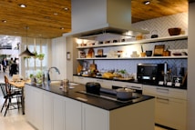 A modern kitchen with a central island featuring a sink and culinary tools. The background includes open shelves displaying various kitchenware such as plates, bowls, and teapots. A microwave is placed on the countertop, and there is a stylish black overhead lamp. The area includes some potted plants, adding a touch of greenery. Wood accents on the ceiling and shelves provide a warm, inviting atmosphere.