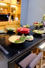A modern kitchen setup featuring a black countertop with various green dishes elegantly arranged. A vibrant red cooking pot sits at the center, surrounded by plates with salads garnished with vegetables. A shelving unit with warm lighting and kitchen appliances can be seen in the background.