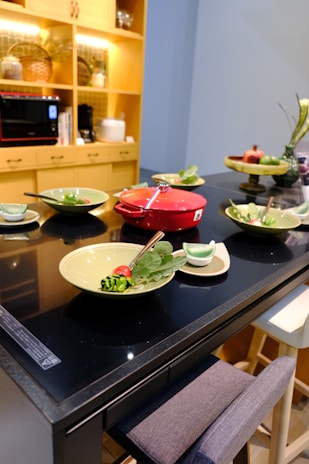 A modern kitchen setup featuring a black countertop with various green dishes elegantly arranged. A vibrant red cooking pot sits at the center, surrounded by plates with salads garnished with vegetables. A shelving unit with warm lighting and kitchen appliances can be seen in the background.