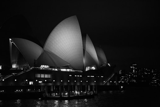 Architectural structure with large, sail-like shapes illuminated against a night sky, with city buildings in the background and reflections on the water below.