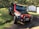 A black and red Jeep with off-road modifications parked on a grassy surface in a wooded area. The vehicle is positioned at an angle, indicating it might have been used for off-road driving. In the background, there are trees and two small metal sheds with some potted plants nearby.