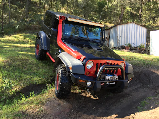 A black and red Jeep with off-road modifications parked on a grassy surface in a wooded area. The vehicle is positioned at an angle, indicating it might have been used for off-road driving. In the background, there are trees and two small metal sheds with some potted plants nearby.