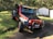 A black and red Jeep with off-road modifications parked on a grassy surface in a wooded area. The vehicle is positioned at an angle, indicating it might have been used for off-road driving. In the background, there are trees and two small metal sheds with some potted plants nearby.