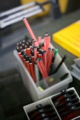 A collection of red and black striped pens is neatly placed in a transparent container. Next to it, a yellow book and some tabs with numbers appear in an adjacent holder.