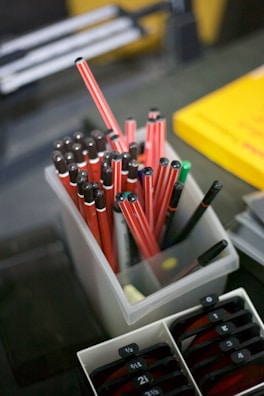 A collection of red and black striped pens is neatly placed in a transparent container. Next to it, a yellow book and some tabs with numbers appear in an adjacent holder.