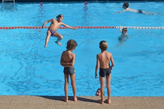 Children are playing in an outdoor swimming pool. One child is jumping into the pool while two others stand at the poolside observing. In the background, other children are swimming and enjoying the water.