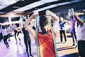 A group of people engage in a fitness class, using resistance bands for exercise. The person in the foreground wearing a red shirt is stretching with a green band, and others in the background mirror the activity in various exercise poses. The setting resembles an indoor gym or fitness center with ample lighting.