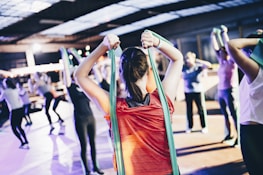 A group of people engage in a fitness class, using resistance bands for exercise. The person in the foreground wearing a red shirt is stretching with a green band, and others in the background mirror the activity in various exercise poses. The setting resembles an indoor gym or fitness center with ample lighting.