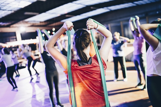 A group of people engage in a fitness class, using resistance bands for exercise. The person in the foreground wearing a red shirt is stretching with a green band, and others in the background mirror the activity in various exercise poses. The setting resembles an indoor gym or fitness center with ample lighting.