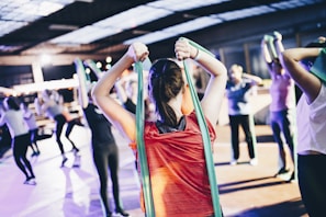 A group of people engage in a fitness class, using resistance bands for exercise. The person in the foreground wearing a red shirt is stretching with a green band, and others in the background mirror the activity in various exercise poses. The setting resembles an indoor gym or fitness center with ample lighting.