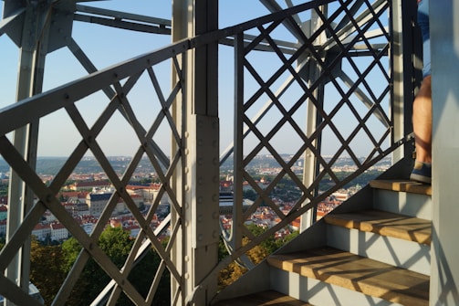 Iron lattice structure overlooking a cityscape, with a person ascending wooden steps on the right. The background features a clear blue sky and distant urban buildings.