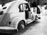 A close-up view of a handcrafted wooden toy car sits on a textured surface. The car features distinct wheels and visible door handles, with a simple and classic design. In the blurred background, there are objects resembling glassware, adding depth to the setting.