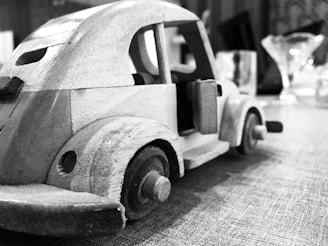 A close-up view of a handcrafted wooden toy car sits on a textured surface. The car features distinct wheels and visible door handles, with a simple and classic design. In the blurred background, there are objects resembling glassware, adding depth to the setting.