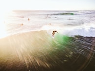 A surfer skillfully rides a wave with several other surfers in the background waiting to catch their turn. The sunlight creates a warm, golden glow across the scene, enhancing the dynamic movement of the water and the energy of the surfers.
