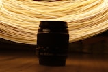 A camera lens is placed centrally against a backdrop of swirling golden light trails. The light creates dynamic patterns that contrast with the dark silhouette of the lens.