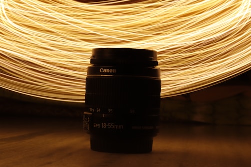 A camera lens is placed centrally against a backdrop of swirling golden light trails. The light creates dynamic patterns that contrast with the dark silhouette of the lens.