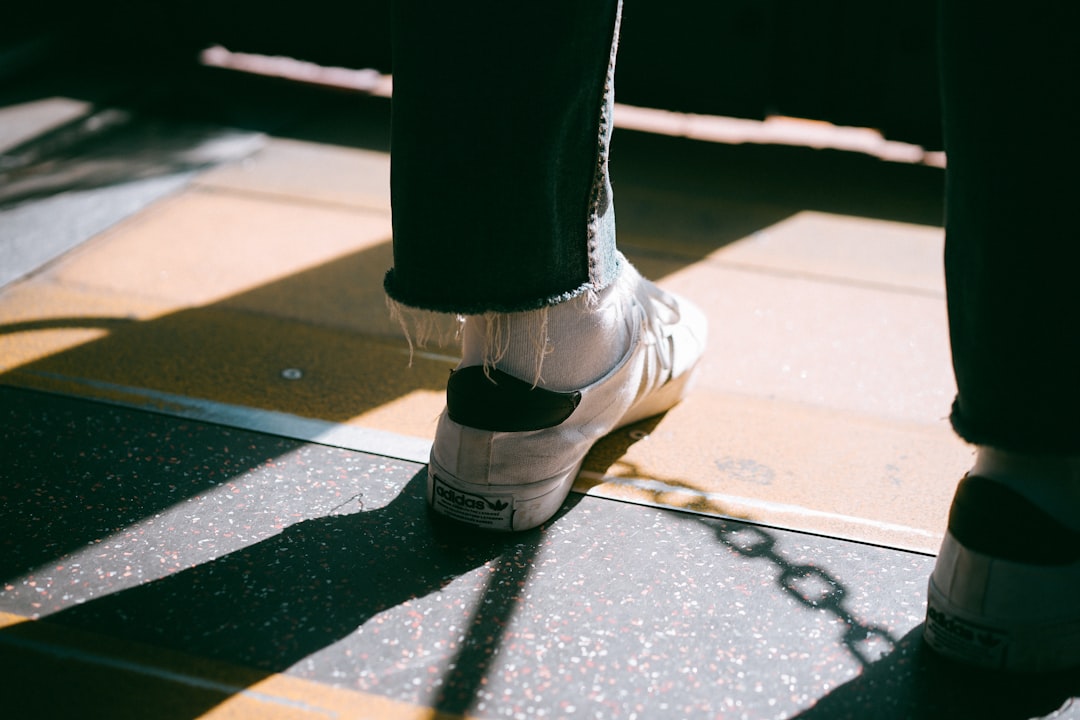 person wearing white low-top sneakers standing on brown wooden floor room