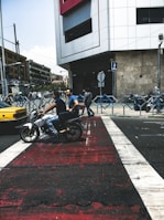 A busy urban street scene featuring a motorcyclist with a passenger riding across a crosswalk. A pedestrian crosses the street nearby, while a taxi and other vehicles are visible in the background. A modern building with a large window dominates the background, and the street is lined with traffic signs and barriers.