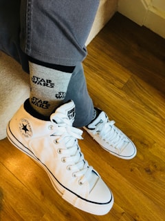 A pair of white Converse sneakers is worn with gray Star Wars-themed socks. The person is dressed in gray pants, sitting or kneeling on a wooden floor.