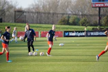 Individuals are engaged in a soccer training session on a grassy field. Some are dribbling soccer balls while a coach oversees the activities. In the background, there is a fence and a sign with the words 'Wash Spirit'.