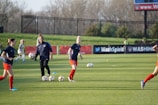 Individuals are engaged in a soccer training session on a grassy field. Some are dribbling soccer balls while a coach oversees the activities. In the background, there is a fence and a sign with the words 'Wash Spirit'.