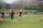 Individuals are engaged in a soccer training session on a grassy field. Some are dribbling soccer balls while a coach oversees the activities. In the background, there is a fence and a sign with the words 'Wash Spirit'.