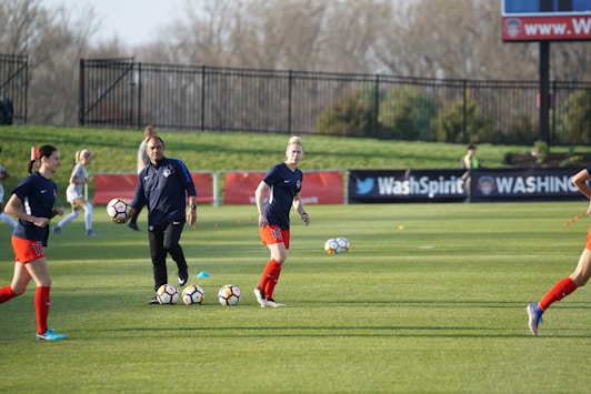 Individuals are engaged in a soccer training session on a grassy field. Some are dribbling soccer balls while a coach oversees the activities. In the background, there is a fence and a sign with the words 'Wash Spirit'.