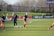 Individuals are engaged in a soccer training session on a grassy field. Some are dribbling soccer balls while a coach oversees the activities. In the background, there is a fence and a sign with the words 'Wash Spirit'.