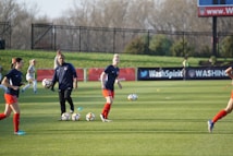 Individuals are engaged in a soccer training session on a grassy field. Some are dribbling soccer balls while a coach oversees the activities. In the background, there is a fence and a sign with the words 'Wash Spirit'.