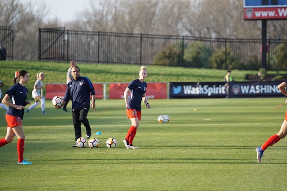 Individuals are engaged in a soccer training session on a grassy field. Some are dribbling soccer balls while a coach oversees the activities. In the background, there is a fence and a sign with the words 'Wash Spirit'.