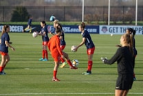 A group of women soccer players are practicing on a grassy field, wearing navy blue tops and red shorts. Some players are actively handling soccer balls, preparing to pass or kick. The field is surrounded by a fence, and there are advertisements visible in the background.