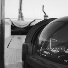 A close-up view of a nautical compass mounted on a boat's deck, with blurred background elements suggesting the boat's structure. The compass's markings are clearly visible, and the image is in grayscale, enhancing its timeless and classic feel.