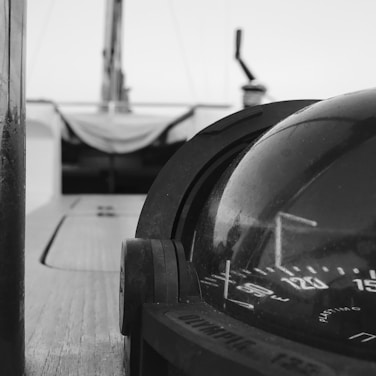 A close-up view of a nautical compass mounted on a boat's deck, with blurred background elements suggesting the boat's structure. The compass's markings are clearly visible, and the image is in grayscale, enhancing its timeless and classic feel.