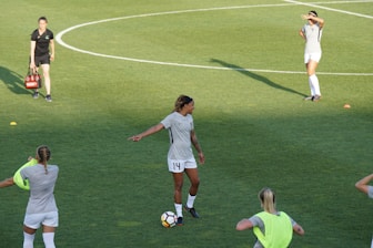 Athletes are on a soccer field engaging in practice. A player wearing a jersey with the number 14 is standing with a soccer ball at their feet, pointing to the right. Teammates are scattered around the field, some stretching or preparing for drills. A coach or staff member is carrying equipment, possibly looking towards the players.