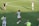 Athletes are on a soccer field engaging in practice. A player wearing a jersey with the number 14 is standing with a soccer ball at their feet, pointing to the right. Teammates are scattered around the field, some stretching or preparing for drills. A coach or staff member is carrying equipment, possibly looking towards the players.