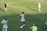 Athletes are on a soccer field engaging in practice. A player wearing a jersey with the number 14 is standing with a soccer ball at their feet, pointing to the right. Teammates are scattered around the field, some stretching or preparing for drills. A coach or staff member is carrying equipment, possibly looking towards the players.