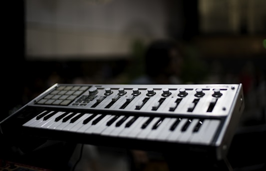 A music keyboard with various control knobs and pads is prominently featured against a blurred background. The keyboard has white and black keys along with several buttons for sound manipulation.