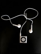 A pair of white earphones connected to a small, square, metallic music player with a circular control pad. The earphones and player are set against a dark background.