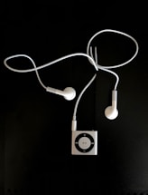 A pair of white earphones connected to a small, square, metallic music player with a circular control pad. The earphones and player are set against a dark background.