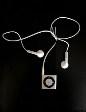 A pair of white earphones connected to a small, square, metallic music player with a circular control pad. The earphones and player are set against a dark background.