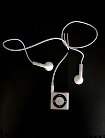 A pair of white earphones connected to a small, square, metallic music player with a circular control pad. The earphones and player are set against a dark background.