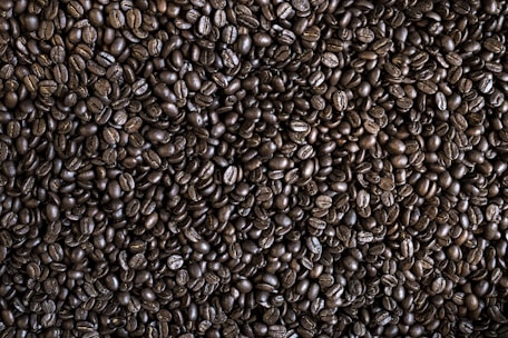 A close-up view of numerous dark roasted coffee beans filling the image. The beans are tightly packed and exhibit a glossy, oily surface characteristic of freshly roasted coffee. The irregular shapes and natural texture of the beans are clearly visible.